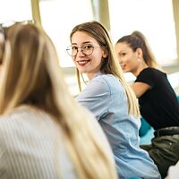 Eine junge Frau mit Brille sitzt im Klassenzimmer und lächelt selbstbewusst in die Kamera.