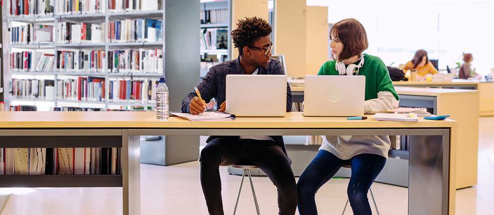 Zwei junge Menschen mit Laptops in einer Bibliothek