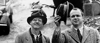 Image du film en noir et blanc : deux hommes à chapeaux côte à côte ; l'homme à droite soulève son chapeau