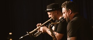 Zu sehen sind zwei Musiker, die ihre Blasinstrumente spielen.