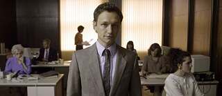 Man in a grey suit in the middle of the frame, behind him, a little blurry, people sitting at desks