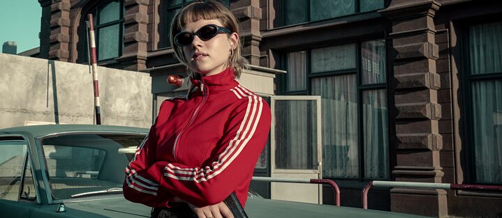 Key art from the Netflix original series “Kleo” shownig the title character portrait by Jella Haase in a red tracksuit jacket holding a pistol