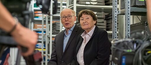 An elderly woman and an elderly man are sitting among shelves with boxes that contain film reels. In front of them are the hands of two people holding cameras.