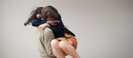 In a side view we see a man in a beige pullover lifting a woman holding her below the knees and behind her back. The woman is hugging him around the neck. Their faces are not visible.