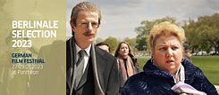 Rabiye Kurnaz vs. George W. Bush – Film Still: Takım elbiseli uzun boylu bir erkek ve mavi paltolu daha kısa boylu bir kadın hayretle bakıyorlar. Arkalarında bir grup insan var ve en arkada Beyaz Saray görünüyor.
