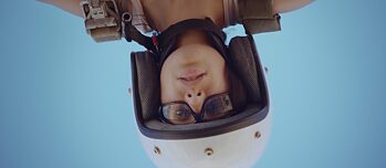 The head of a young woman wearing a helmet, glasses and a rucksack is turned upside down in the photo. The background shows a clear blue sky.