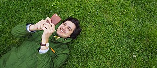Ein lächelnder junger Mann liegt auf dem Rücken im Gras und hält ein Smartphone in den Händen. Raum für redaktionellen Text. Bildserie für die Bewerbung der Sprachkurse und Prüfungen in der Region SAM.