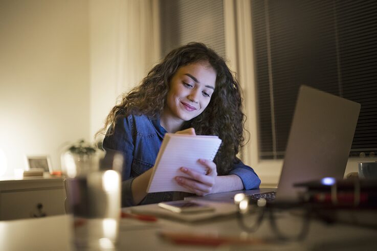 junge schwarzhaarige Studentin studiert zu Hause mit Laptop und macht sich Notizen im Notizbuch.