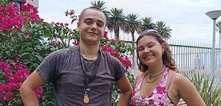 Ein junger Mann und eine junge Frau stehen in einem Garten und schauen lächelnd in die Kamera. Im Hintergrund stehen Palmen und ein blühender Busch.