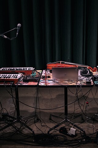 Imagen frontal de escenario, sobre un par de mesas se ve un violín, un laptop y varios teclados