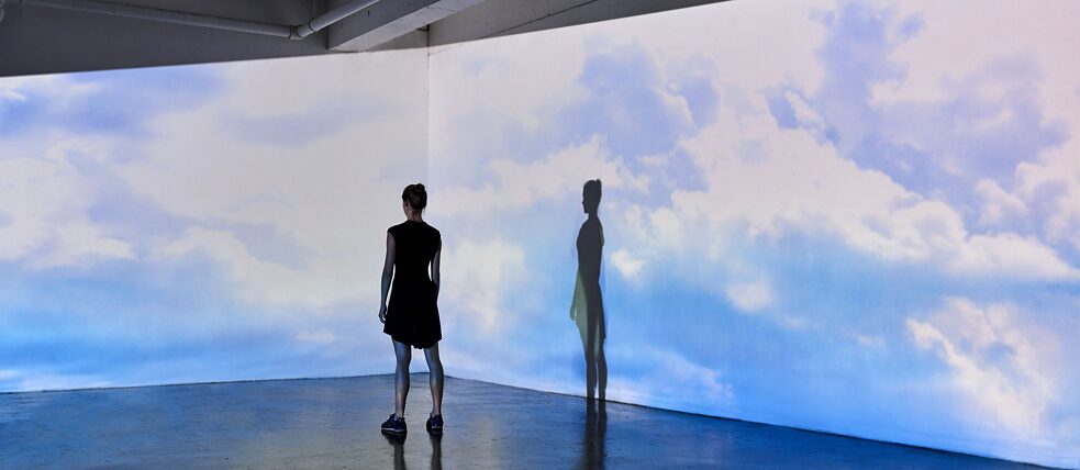 Una mujer joven está de pie en una galería y observa las paredes sobre las que se proyectan el cielo y las nubes.