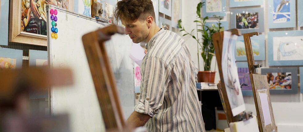 Чоловік пише на білій дошці, на задньому плані художня майстерня, до розвішані і розставлені картини