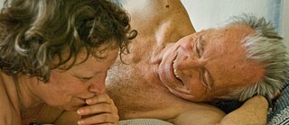 Deux personnes âgées couchées torses nus sur un lit, sourire aux lèvres