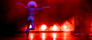 A blue lit person from back perspective dancing in front of a red (fiery) background.