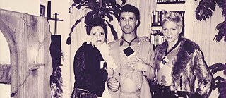 Drei Personen in Kostümen, die vor einer Wohnzimmerkulisse für die Kamera posieren.