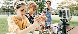 Drei junge Leute bauen im Freien einen Mini-Roboter. Einer von ihnen zeichnet den Vorgang mit seinem Handy auf