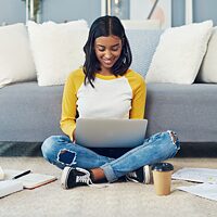 Eine junge Frau in modischen Jeans sitzt im Wohnzimmer auf dem Boden vor einer Couch und blickt lächeln auf ein Laptop.