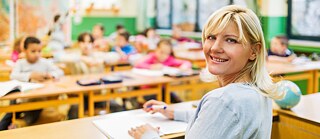 Una docente mira a la cámara en primer plano, al fondo de la imagen se ve una sala de clases llena de alumnos/as