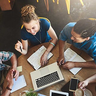 View from above of young people in a café. On the table are laptop, smartphone and opened notebooks.