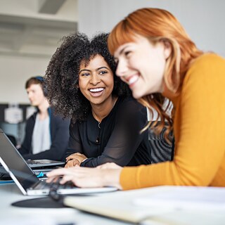 Dos mujeres jóvenes y sonrientes están sentadas en una oficina frente al escritorio. Delante de ellas se encuentran varias pantallas y una computadora portátil. 