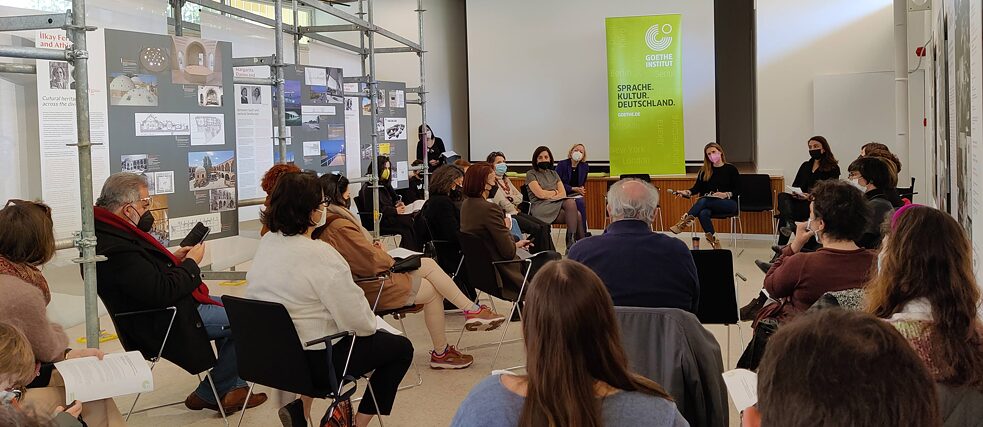 Περίπου είκοσι άτομα, τα περισσότερα εκ των οποίων γυναίκες, κάθονται σε κυκλική διάταξη σε έναν εκθεσιακό χώρο, όπου υπάρχουν πίνακίδες προσαρμοσμένοι στους τοίχους και σε μια κατασκευή σκαλωσιάς.