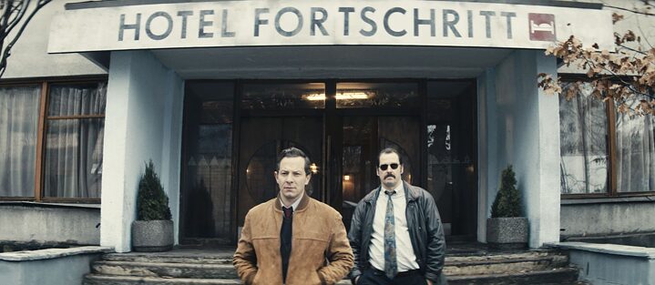 Szene aus dem Film „Freies Land“. Zwei Männer stehen vor dem Eingang des Hotels Fortschritt.
