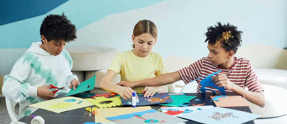 two boys and a girl sit at a table and cut out pieces of paper 