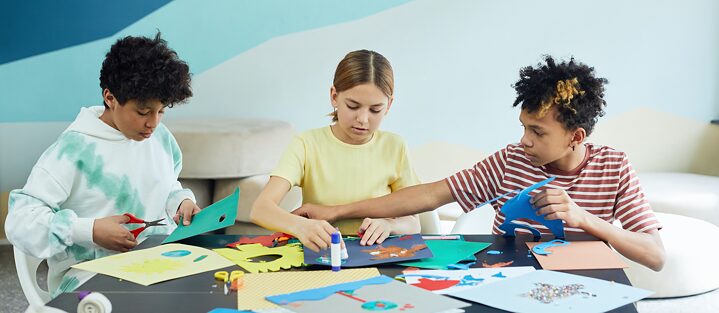 two boys and a girl are sitting at a table and cut out pieces of paper 