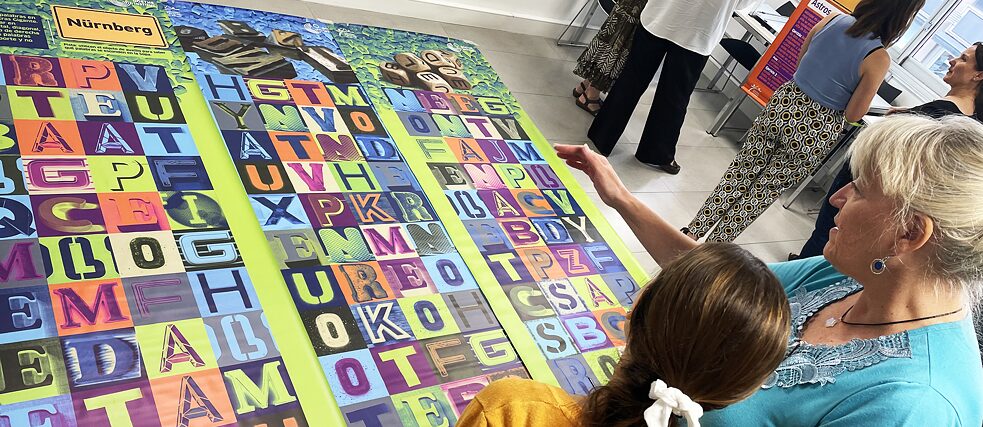Imagen de una profesora y una alumna paradas frente a la estación de juego de Nürenberg, que consiste en unos afiches que forman una sopa de letras