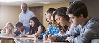 Schüler während einer Prüfung im Klassenraum. Der Lehrer steht beobachtend im Hintergrund. © © Goethe-Institut, Getty Images Beratung und Information Deutschprüfungen