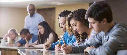 Schüler während einer Prüfung im Klassenraum. Der Lehrer steht beobachtend im Hintergrund.