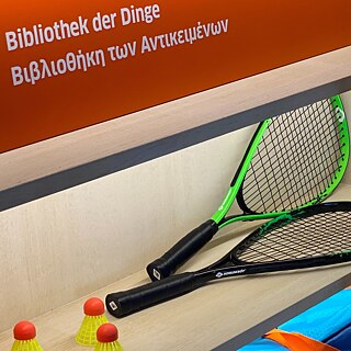 Ein Regal mit verschiedenen Gegenständen, u.a. ein Tennisschläger und ein rotes Schild "Bibliothek der Dinge"