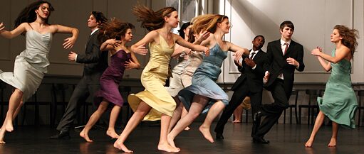 Fotoğrafta dans eden gençler görülüyor. Erkekler koyu renk takım elbise, kızlar ise rengarenk elbiseler giymiş. 