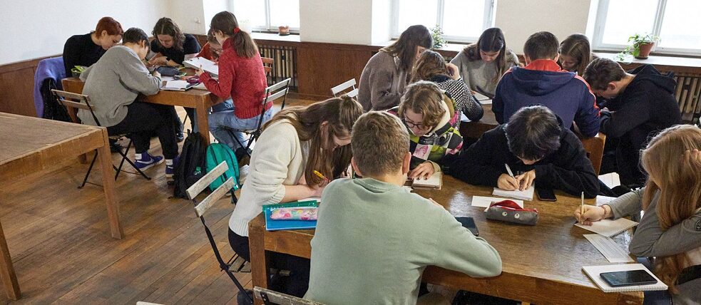 Žáci sedí u stolů a píší.