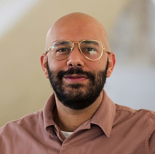 A man with a shaved head, beard and myopic glasses looks smilingly into the camera (Evagoras Vanezis). He wears jeans and a brown jacket and rests his left elbow on a white pillar.