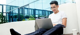 Junger Mann sitzt mit einem Laptop vor einem Firmengebäude