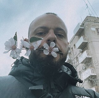 A man with a beard (Stelios Kallinikou), a black jacket and a serious look is holding an almond branch with his mouth and looking at the camera.