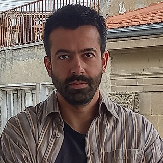 A man with short black hair and beard is calmly looking into the camera with his arms crossed.
