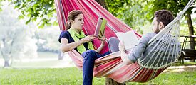 Eine Frau und ein Mann sitzen entspannt in einer Hängematte im Park. Sie liest mit dem Tablet und er liest ein Buch. Neben ihnen liegt ein Bücherstapel auf dem Rasen.