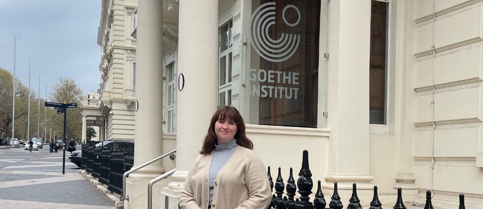 Sarah steht vor dem Institut mit cremefarbenem Pullover