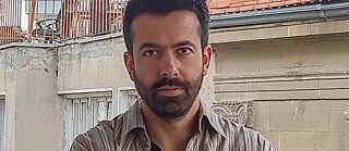 A man with short black hair and beard is calmly looking into the camera with his arms crossed.