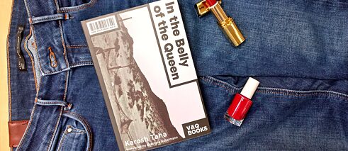 The book 'In the Belly of the Queen' is lying on a pair of jeans together with red lipstick and nail polish.