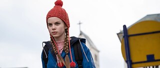 Kırmızı dikişli şapkası, mavi ceketi ve iki uzun at kuyruğu olan bir kız kararlılıkla önüne bakıyor. 