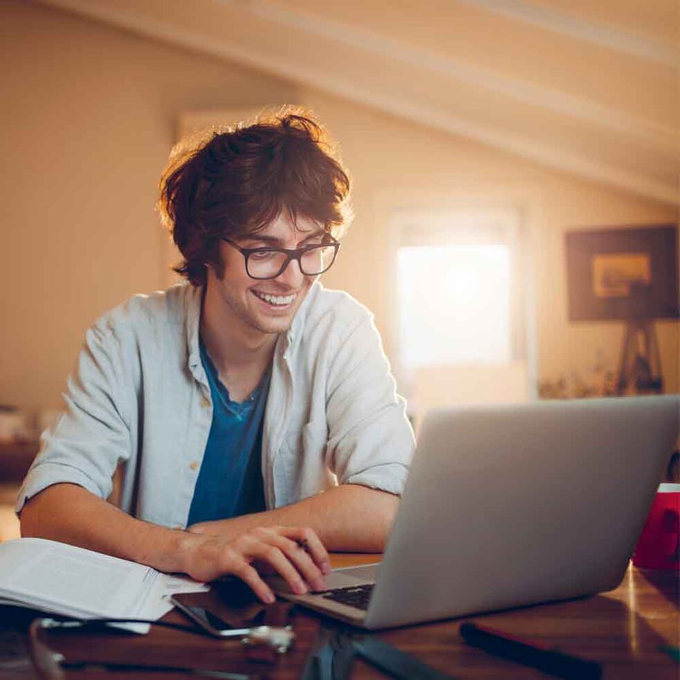 garoto sorri e estuda dentro de casa, sentado em mesa com caderno e computador 