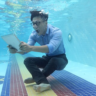  Ein Mann sitzt in Geschäftskleidung am Grunde eines Schwimmbads, unter Wasser. Er blickt erstaunt auf ein Tablet und schreit vor Freude.