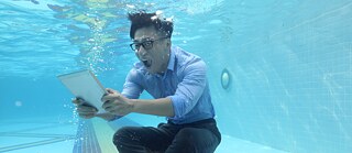  Ein Mann sitzt in Geschäftskleidung am Grunde eines Schwimmbads, unter Wasser. Er blickt erstaunt auf ein Tablet und schreit vor Freude.