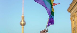 Ein Fernsehturm und eine Regenbogenfarben-Flagge vor himmelblauem Hintergrund 