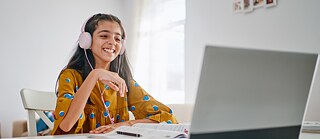Ein Mädchen sitzt mit Buch und Kopfhörern vor dem Computer und starrt lächelnd auf den Bildschirm