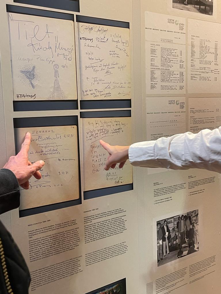 Besucher*innen der Ausstellung im Goethe-Institut Athen vor dem Installationspanel mit Seiten aus den Gästebüchern der 60-Jahre mit Unterschriften und Kommentaren von bekannten Künstler*innen und Schriftsteller*innen der Zeit in der Bibliothek 