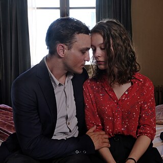 "Transit" filminden bir sahne: Takım elbiseli bir adam ve kırmızı bluzlu bir kadın bir yatakta yan yana ve yüzleri birbirine dönük olarak oturuyor. Arkadaki oda bir otel odası gibi görünüyor.